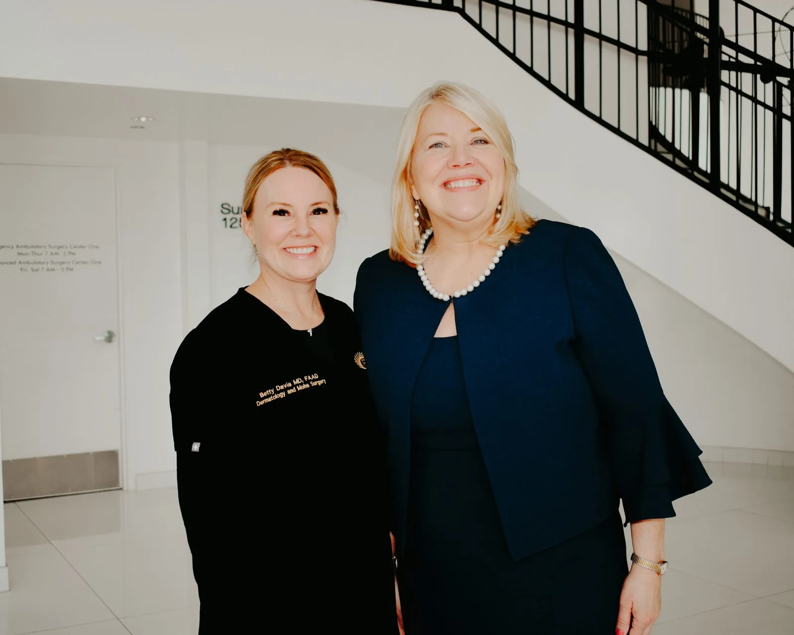 Two women smiling in a modern clinical hallway with a staircase in the background